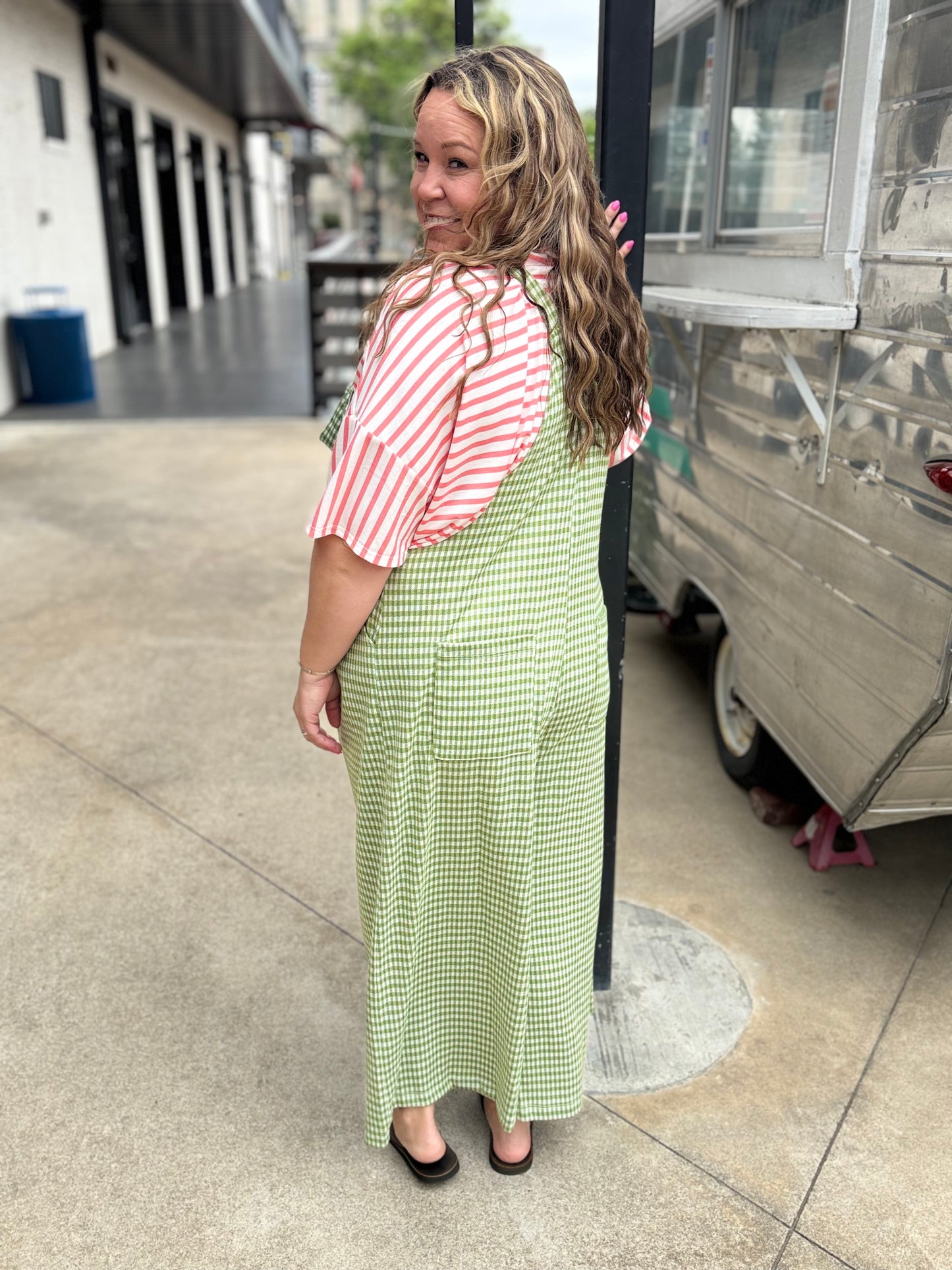 Woman in a pink striped shirt and green checkered dress standing on a sidewalk next to a food truck.
