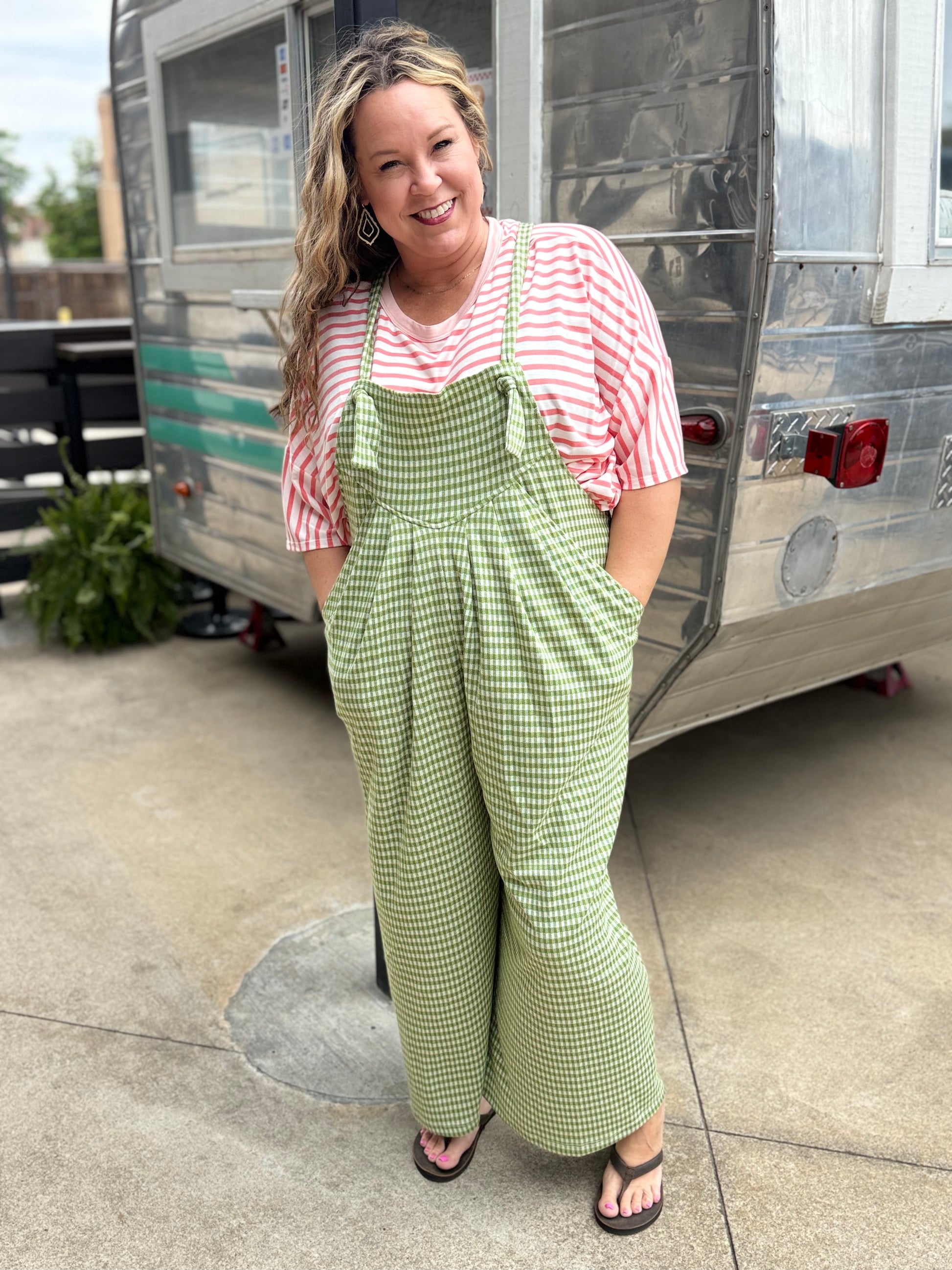 Woman wearing a red and white striped shirt and green checkered overalls standing in front of a vintage trailer.